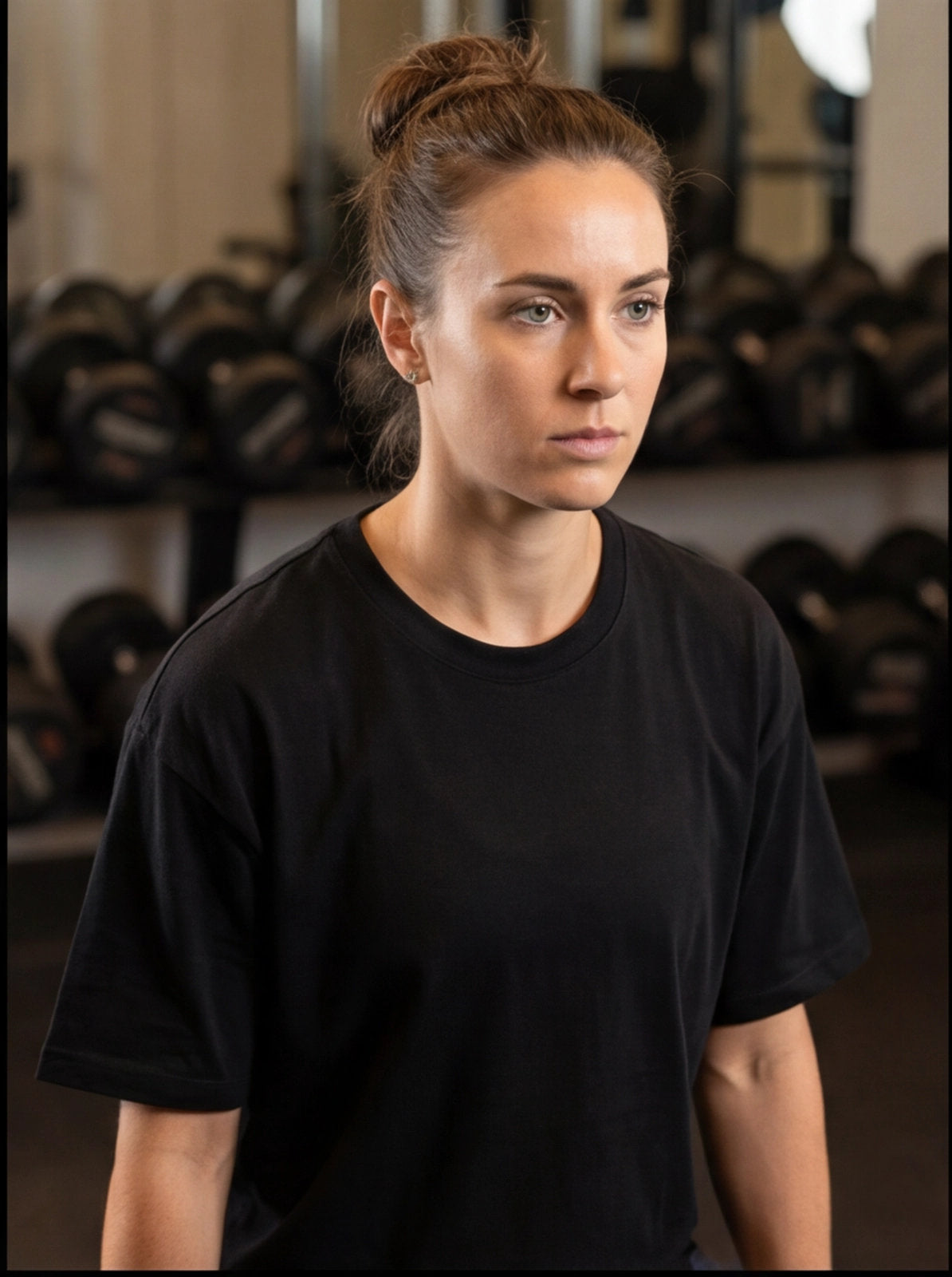 Woman in black fitness t-shirt at gym, focused expression, dumbbells in background for functional training