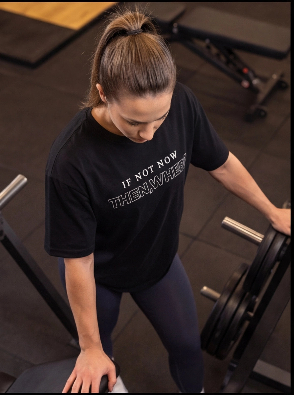 Woman in gym wearing black 'IF NOT NOW THEN, WHERE?' t-shirt, preparing for weightlifting workout