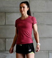 Woman in athletic maroon t-shirt and black workout shorts posing against a concrete wall