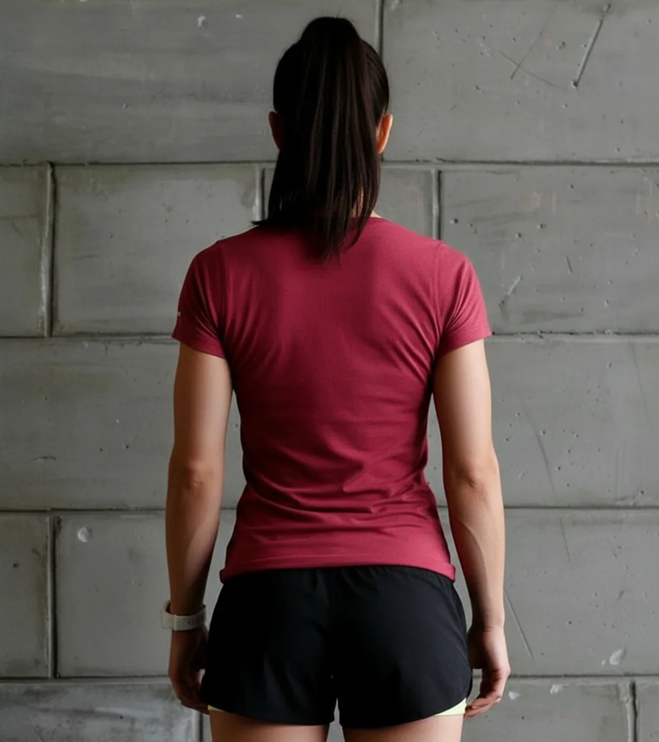 Woman in athletic maroon t-shirt and black shorts facing concrete wall, fitness apparel