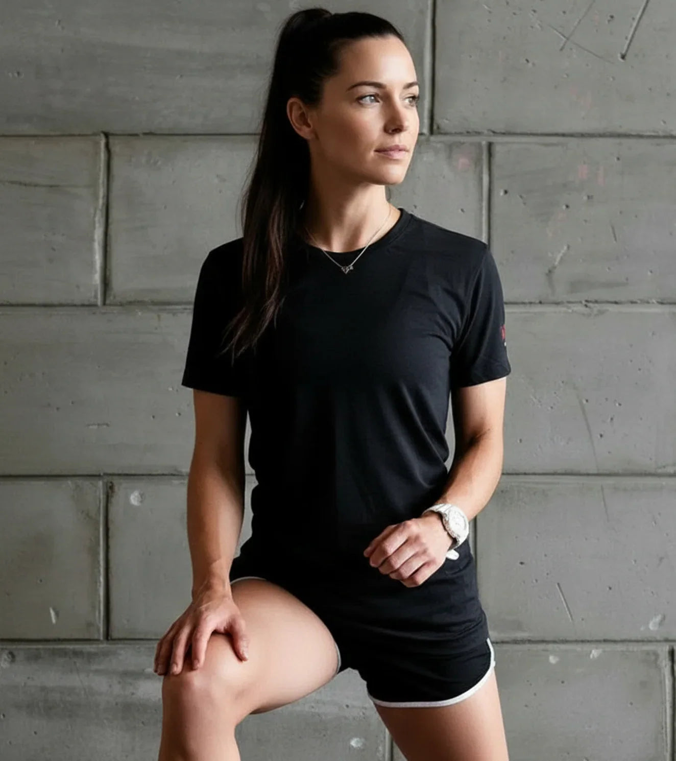 Athletic woman in black workout shirt and shorts posing against concrete gym wall