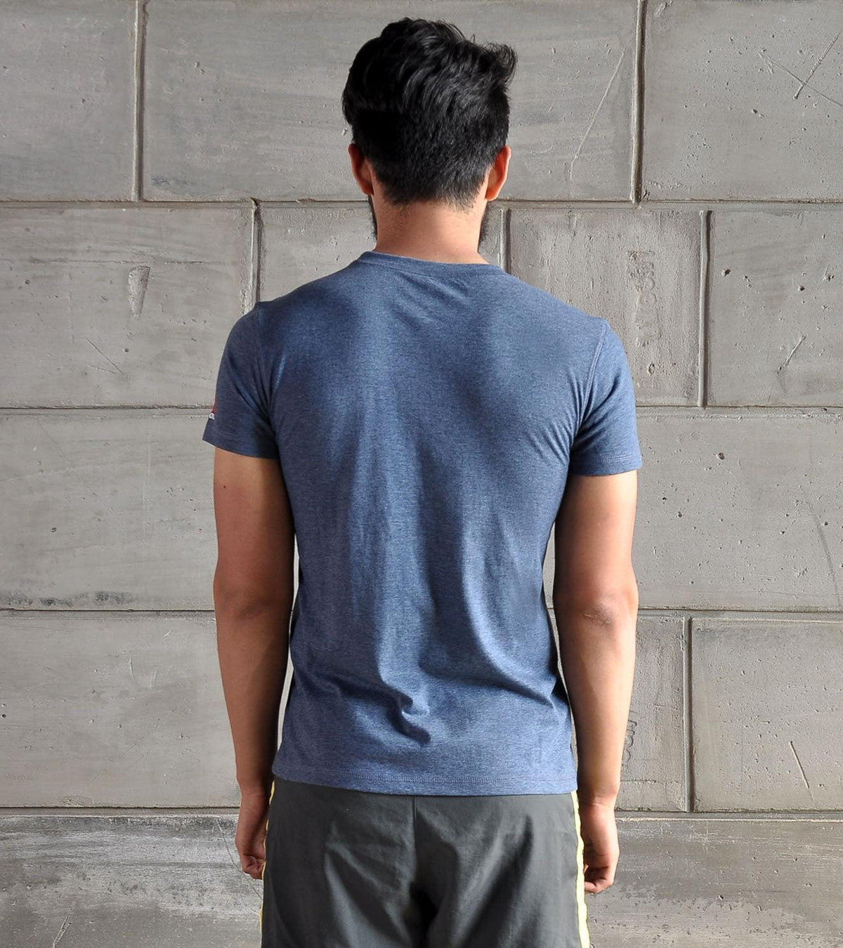 Back view of a man wearing a blue athletic t-shirt and grey gym shorts against a grey wall.
