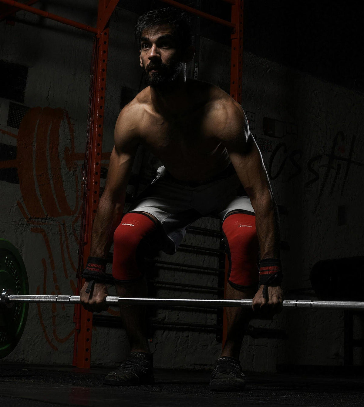 Shirtless athlete wearing knee sleeves and wrist wraps preparing for a barbell lift.