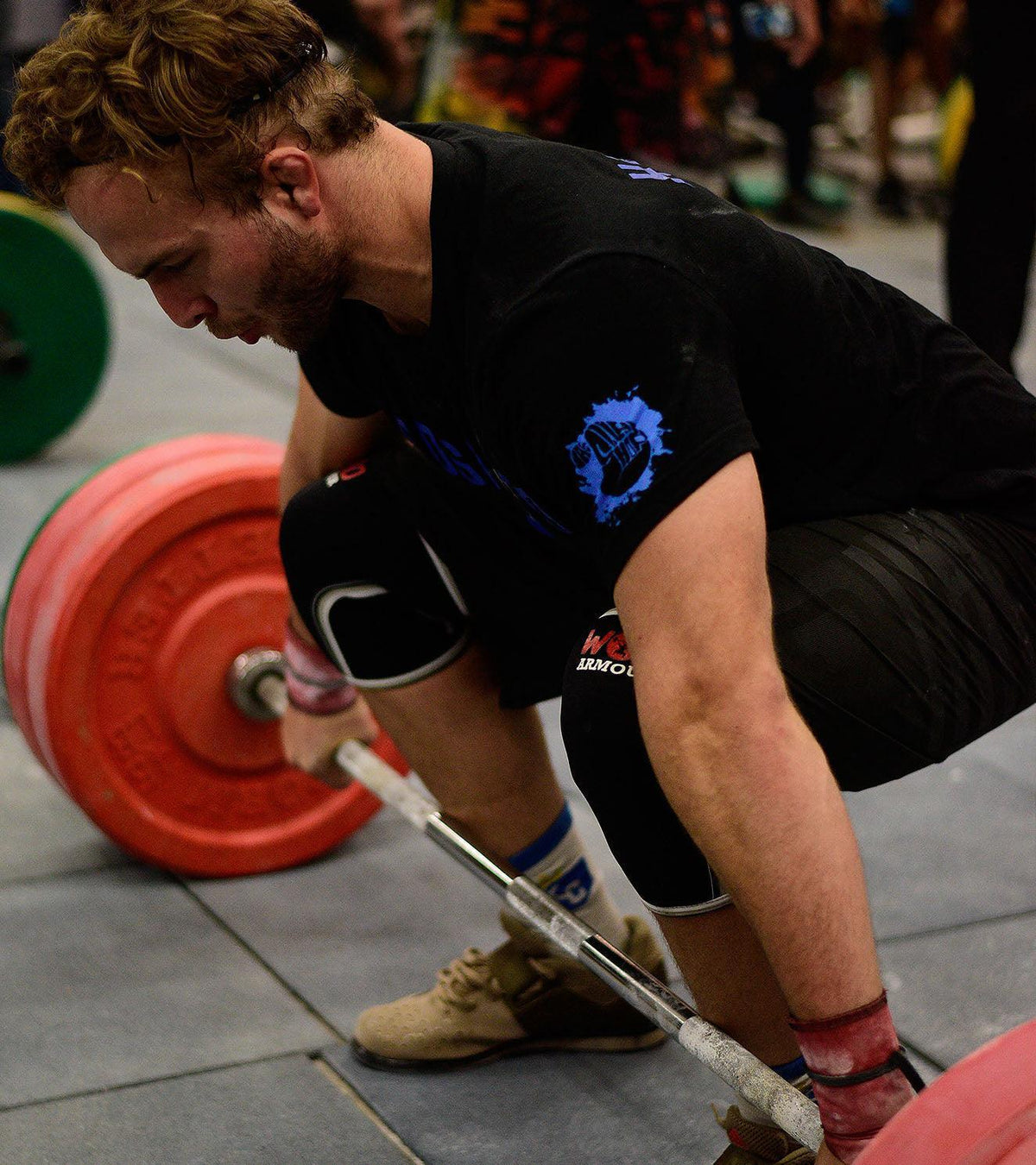 Male athlete wearing knee sleeves and wrist wraps lifting a barbell with red weight plates.