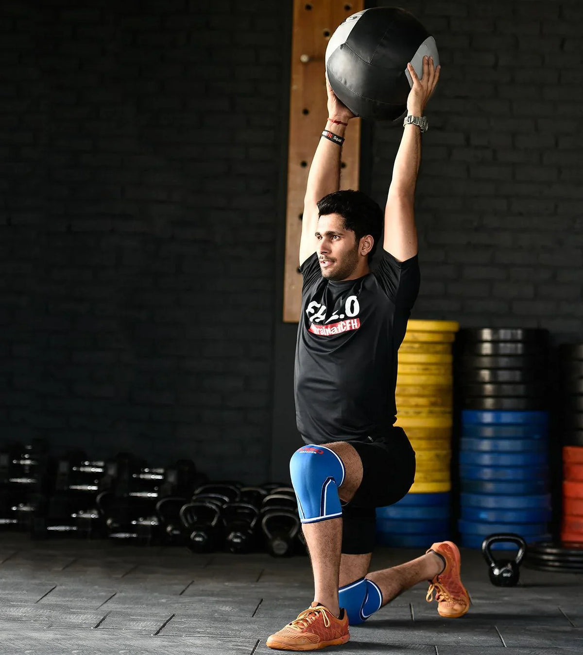 Man in gym wearing blue knee sleeves holding a medicine ball overhead during a workout.